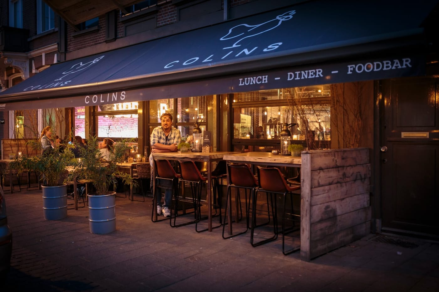 Colin voor de deur van Restaurant Colins in Breda, in de schemer met de zaak vol achter hem