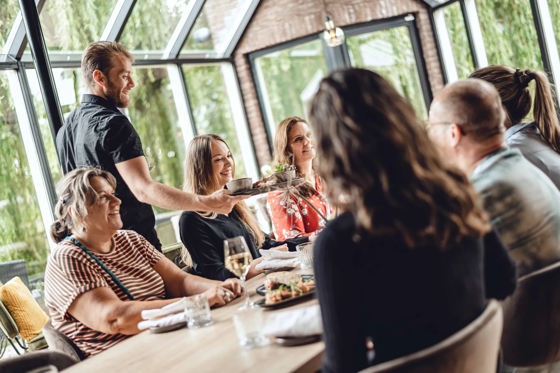 Gasten aan de lange tafel van Villa Kalkoven in Meppel worden bediend, met daglicht door de serrewanden