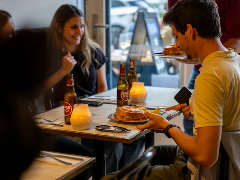 Twee gasten proosten aan tafel bij Portugalia Churrasco in Rotterdam terwijl de bediening een francesinha brengt